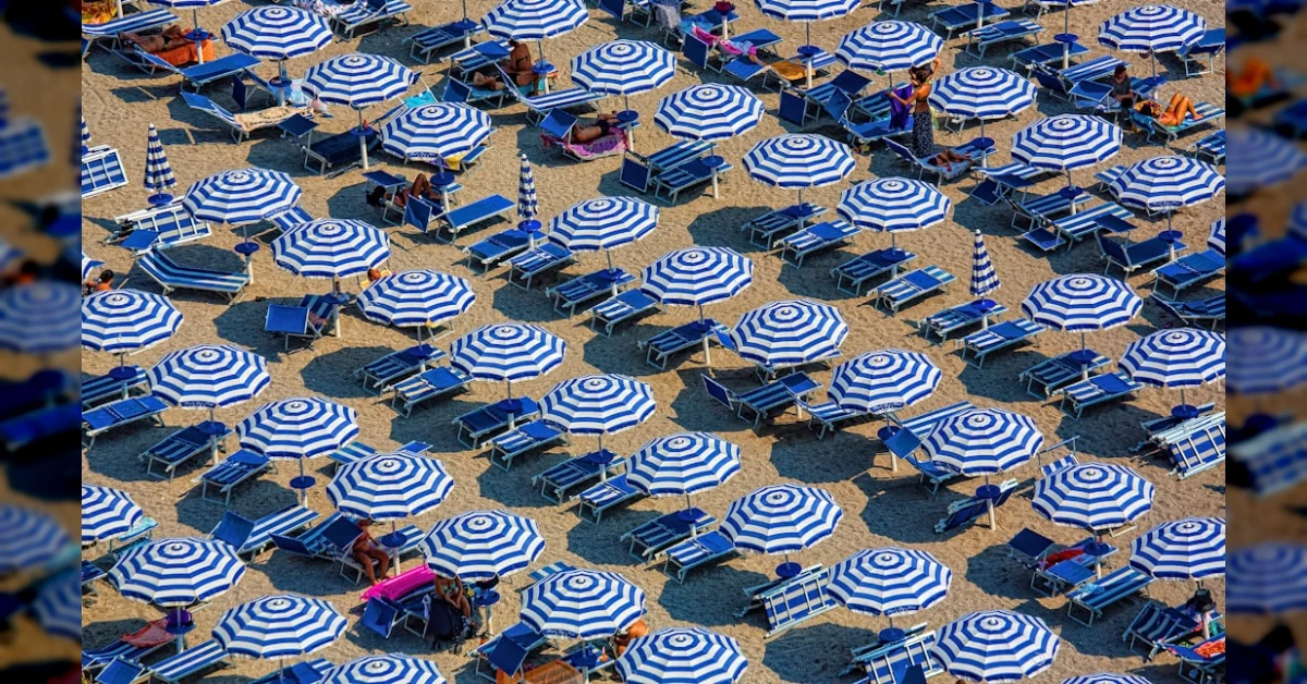 Ombrelloni e sdraio su una spiaggia affollata, con ombre proiettate in diverse direzioni dal sole.