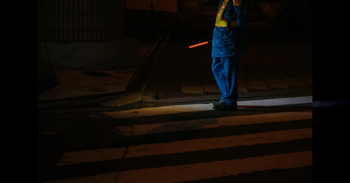Pedone con giubbotto riflettente di notte su una strada extraurbana, con fari di un'auto in avvicinamento.