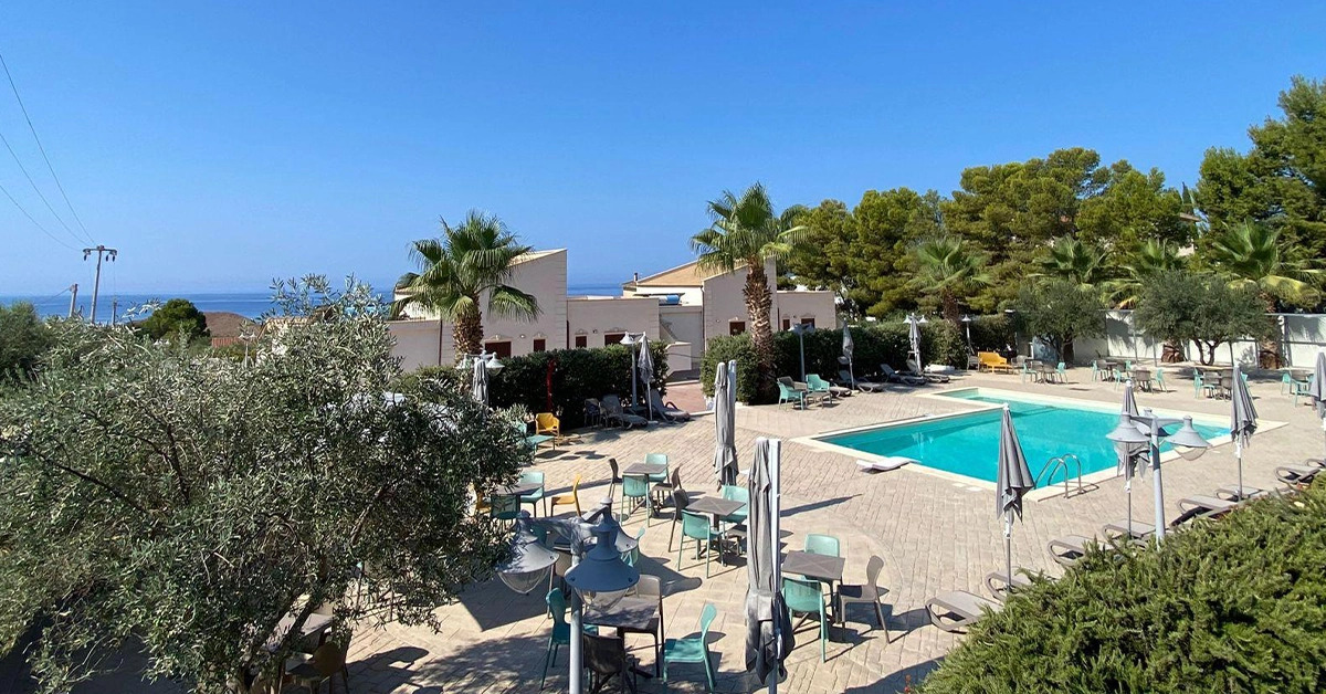 Veduta panoramica di Giotera Village a Sciacca, con l'area piscina, i tavoli all'aperto e il mare sullo sfondo.